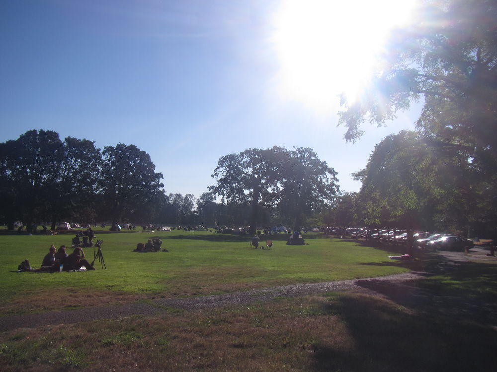 A sunny day at a park.  The sky is blue; the sun is shining; there are a few groups of people, one of which has a camera on a tripod; there are a few trees as well.