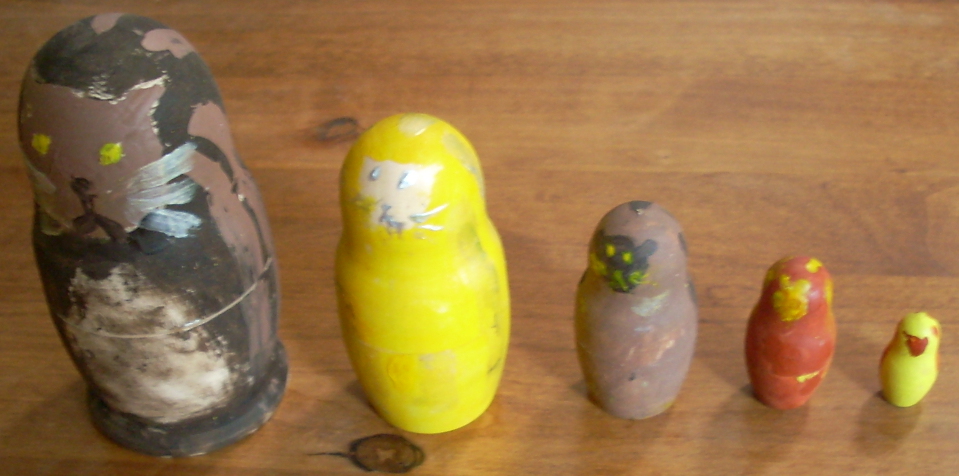 A series of five nesting dolls, painted to have cat faces.  The left (largest) is black with brown stripes and a brown face and a white tummy.  The next one is yellow with a whitish face; then brown with a black face and black stripes; then red with a yellow face and yellow stripes; then the smallest is yellow with a red face.