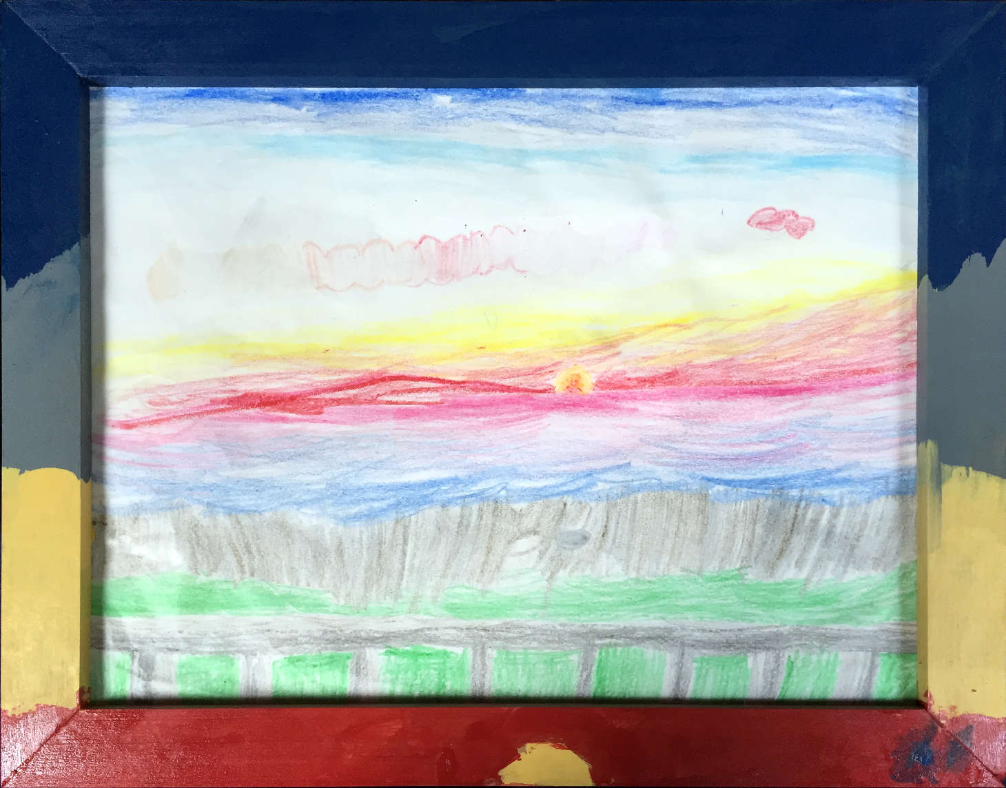 A sunset.  The sky is red, yellow, white, cyan, and dark blue.  There's a long, horizontal cloud, with many colors, yellow on the left, then orange, red, and pink.  There's another pink cloud to the right.  The sun is halfway below the horizon, and is a darkish orange in the middle, and a bright yellow around the outside.  Below the horizon is the ocean, which is magenta, then purple, then blue.  Below that is the beach, with some seashells visible, and then some grass and a rail.  Around the picture is a wooden frame; the top part of the frame is painted dark blue, then below that gray, below that yellow, and at the bottom red.  There's a yellow half-sun painted at the bottom of the frame.